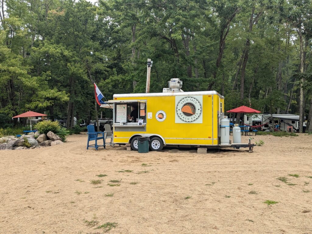 A yellow food truck is parked on a sandy area, surrounded by trees and picnic umbrellas.