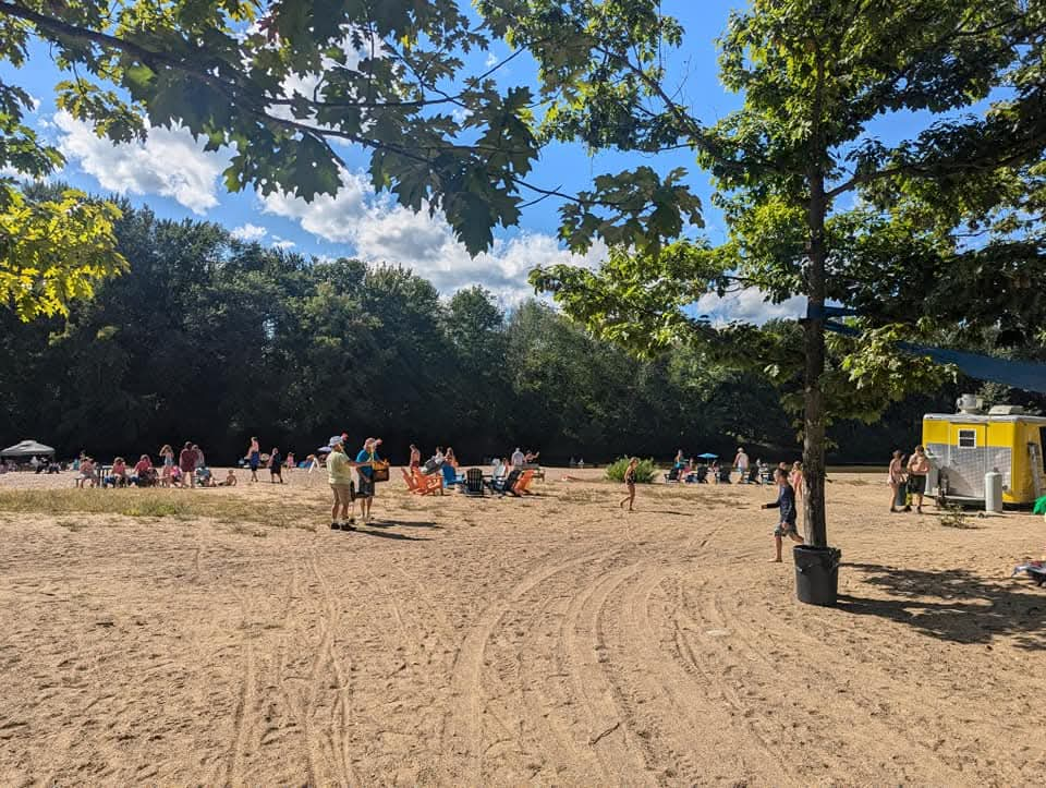 A sunny beach scene with people relaxing, trees, and a food stand near sandy areas.